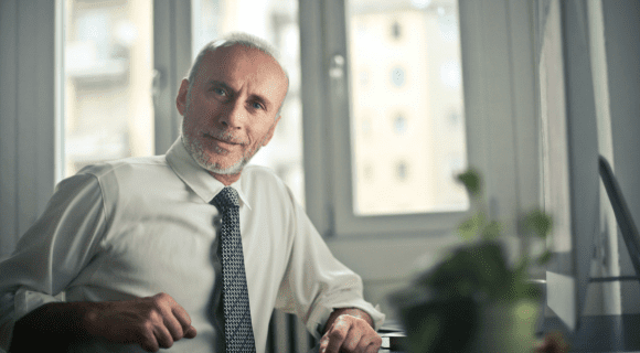 A mature gentleman wearing a shirt and tie looks into the camera while sat in his office.