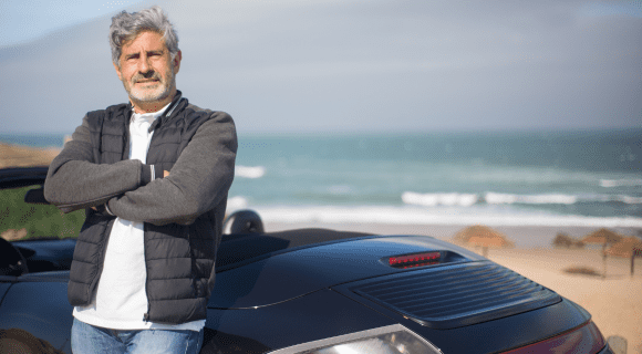 A mature man with greying hair stands in front of a convertible car with a beach in the distance.