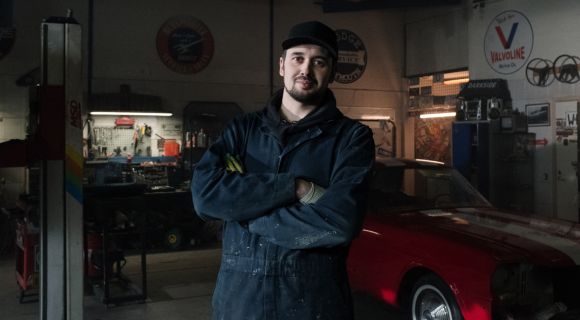 A mechanic wearing blue overalls and a cap, in a garage, which his armed folded, smiles.