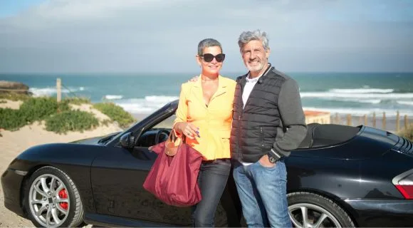 An older couple in front of a convertible car near the beach