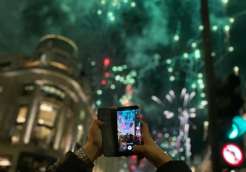 A close-up of hands holding a phone as fireworks explode above them.