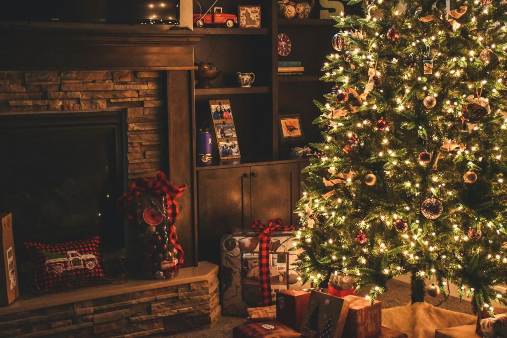  A living room setting decorated with Christmas decorations including presents with red tartan ribbons under a Christmas tree with golden lights and baubles, in front of an unlit fireplace. 