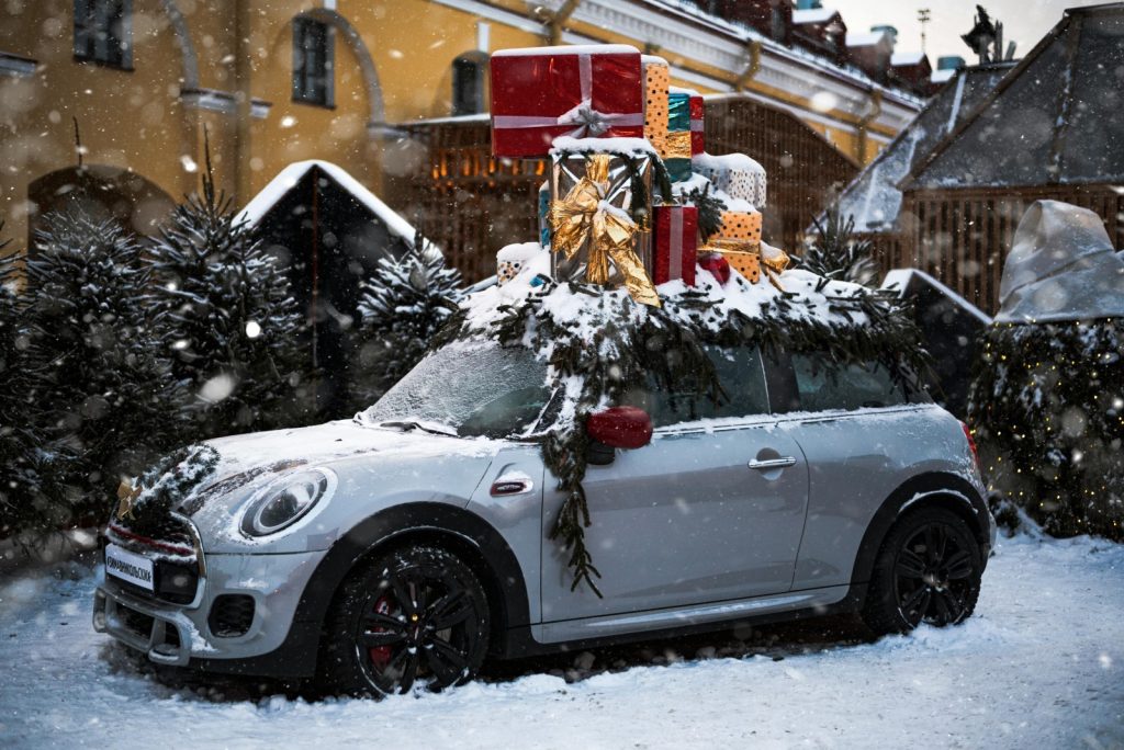 A silver mini in the snow a pile of presents and a Christmas tree on the roof, parked near Christmas trees.