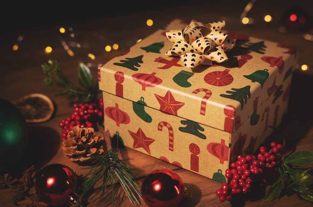 A beige Christmas gift box surrounded by baubles, lights and a wreath. The paper is decorated with a red and green pattern featuring motifs of candy canes, stars, stockings and baubles.  